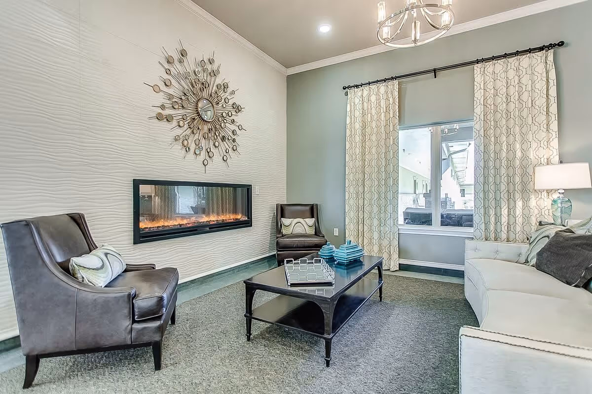 A cozy living room with a modern electric fireplace mounted on a textured white wall. The room features two dark leather armchairs with patterned pillows, a white sofa with multiple cushions, a black coffee table with decorative items, a window with patterned curtains, and a ceiling light fixture.