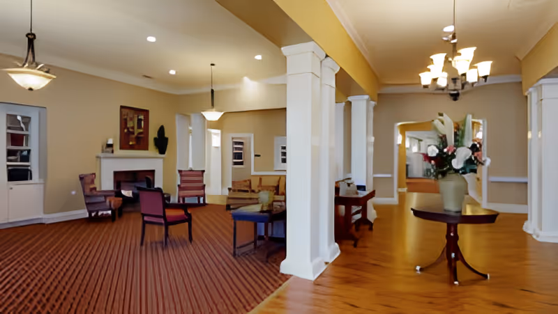 Spacious senior living common room with seating areas, columns, a fireplace, chandeliers, and a center table with flowers.