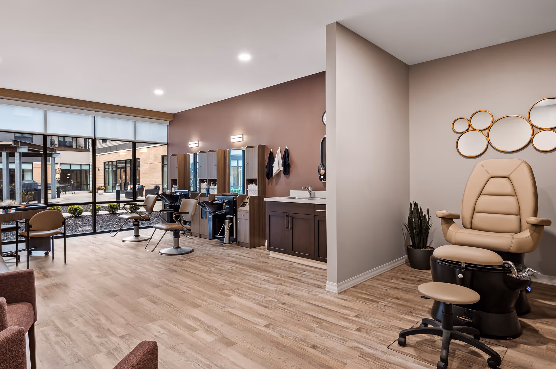 Interior view of a salon area in a senior living facility with salon chairs, wash basins, mirrors, and a pedicure chair. Large windows show an outdoor courtyard with seating and plants.