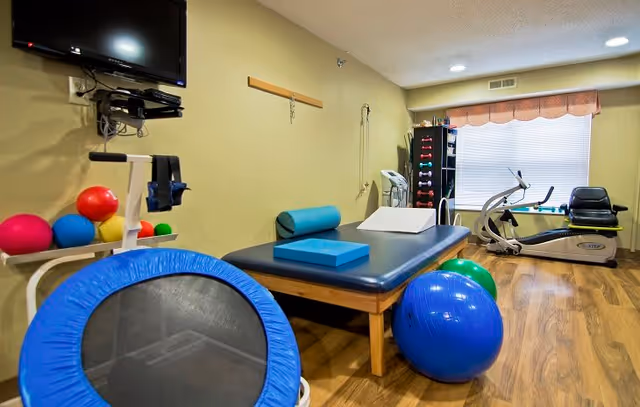 A small exercise room with a blue mini trampoline, colorful exercise balls, a padded exercise table with foam blocks, a stationary recumbent bike, and a rack of dumbbells. The room has wooden flooring, beige walls, a window with blinds and a valance, and a wall-mounted TV.