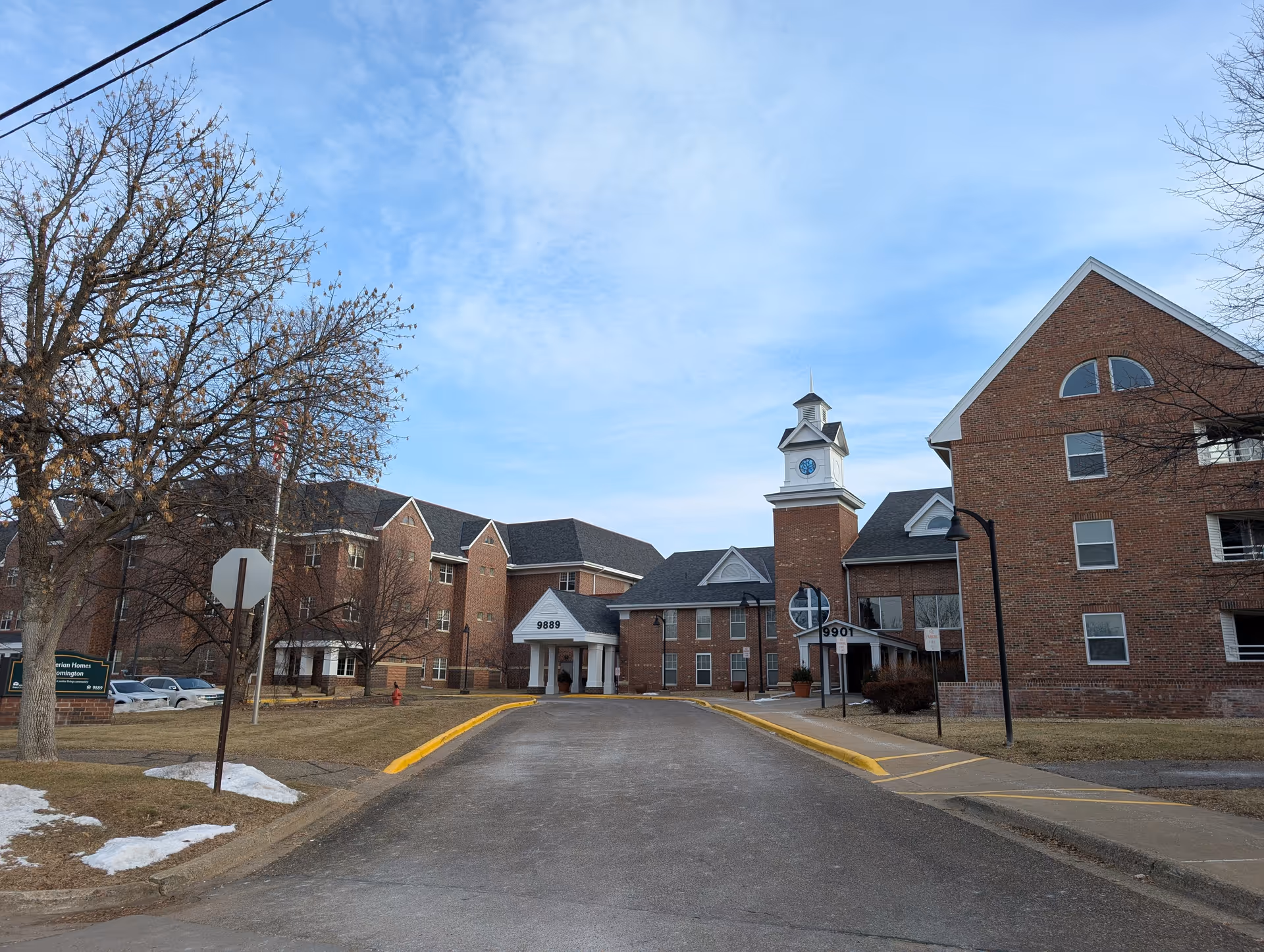 Exterior view of Presbyterian Homes Of Blooming, showing a large brick building with multiple windows and a clock tower. The driveway leads to the main entrance with the numbers 9889 and 9901 visible. Leafless trees and patches of snow are seen on the grass under a partly cloudy sky.