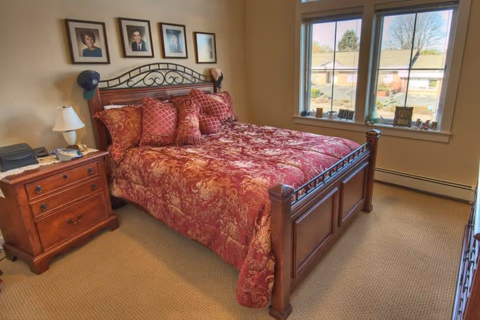 A bedroom with a wooden bed frame and a red patterned bedspread with matching pillows. There is a wooden nightstand with a lamp, phone, and other items next to the bed. Above the bed are four framed photographs on the wall. A large window with a view of neighboring buildings and trees is on the right side of the room.