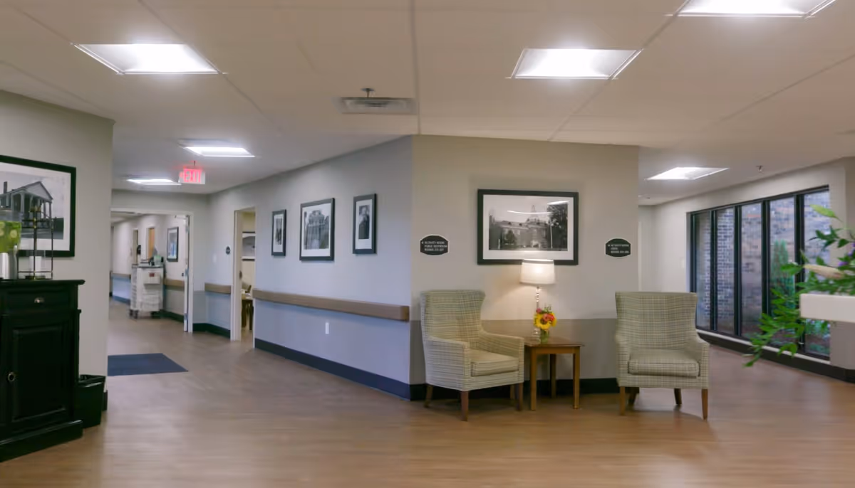 A well-lit hallway in a senior living facility with light-colored walls and wood flooring. There are framed black and white photographs on the walls, two upholstered armchairs with a small wooden table and a lamp between them. Large windows on the right side allow natural light to enter, and a dark wooden cabinet with decorative items is visible on the left.