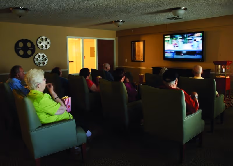 A group of elderly people seated in green armchairs watching a television screen in a dimly lit room. Some are holding popcorn containers. The room has beige walls with decorative film reels on one wall and a popcorn machine in the corner.