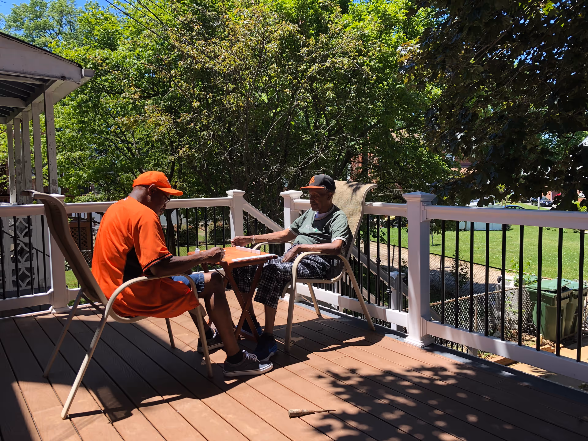 Two elderly men sitting on a wooden deck outdoors, playing a board game on a small table between them. The deck has white railings with black balusters and is surrounded by green trees and grass under a sunny sky.