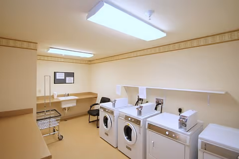 A laundry room with three front-loading washing machines and one dryer lined up against a wall. There is a small sink with a chair next to it, a metal laundry cart, and beige walls with a decorative border near the ceiling. The room is lit by two rectangular fluorescent ceiling lights.