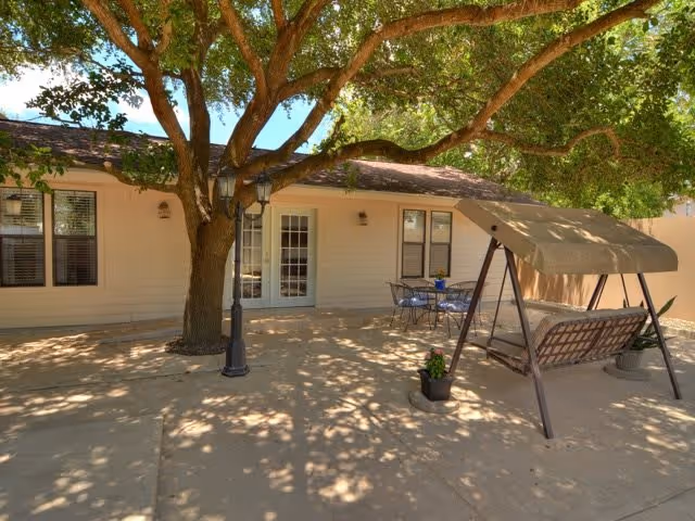 Shaded outdoor patio courtyard with a large tree, a canopy swing, and a table and chairs in front of a building with French doors.