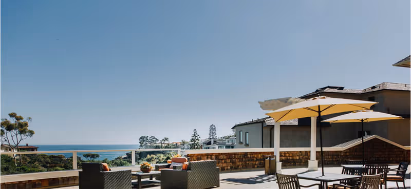 Outdoor patio area at Crown Cove with wicker seating, tables, chairs, and large umbrellas overlooking a scenic view of the ocean and greenery under a clear blue sky.