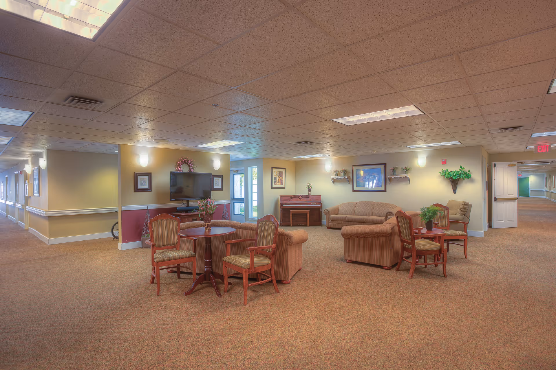 A spacious common area in a senior living facility with beige walls and carpet. The room features multiple seating arrangements including a round table with four chairs, a sofa, armchairs, and a small table with two chairs. A flat-screen TV is mounted on the wall above a small cabinet, and a piano is placed against the far wall. The area is decorated with framed pictures, wall-mounted plants, and a wreath. Hallways extend from both sides of the room.