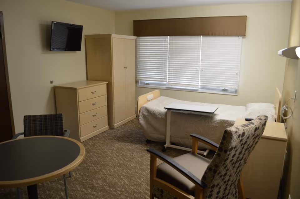 A simple senior care bedroom with a single bed, overbed table, dresser and wardrobe, wall-mounted TV, and a small table with chairs.