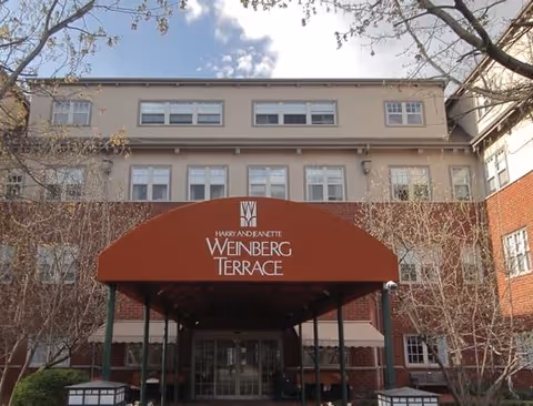 Front exterior view of a multi-story brick building with a red canopy entrance labeled 'Harry and Jeanette Weinberg Terrace'. Leafless trees and a partly cloudy sky are visible.
