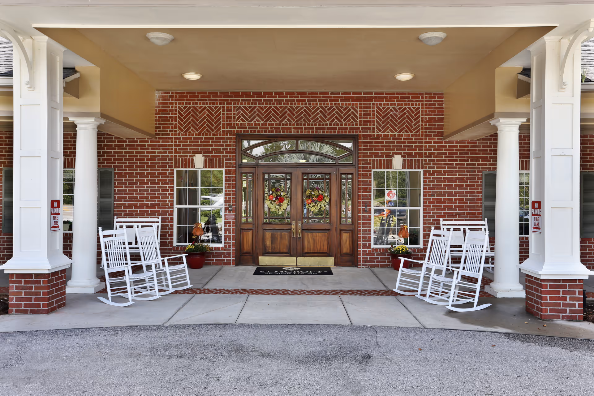 Entrance of a building with a covered porch featuring white columns and four white rocking chairs. The building has a red brick exterior with two windows on either side of a wooden double door decorated with floral wreaths. There are potted plants near the windows and signs indicating no parking in the fire lane.