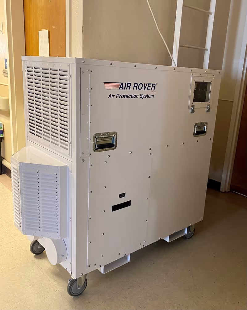 A large white portable air filtration unit labeled 'AIR ROVER Air Protection System' on wheels, placed indoors on a tiled floor near a wooden door and a wall.