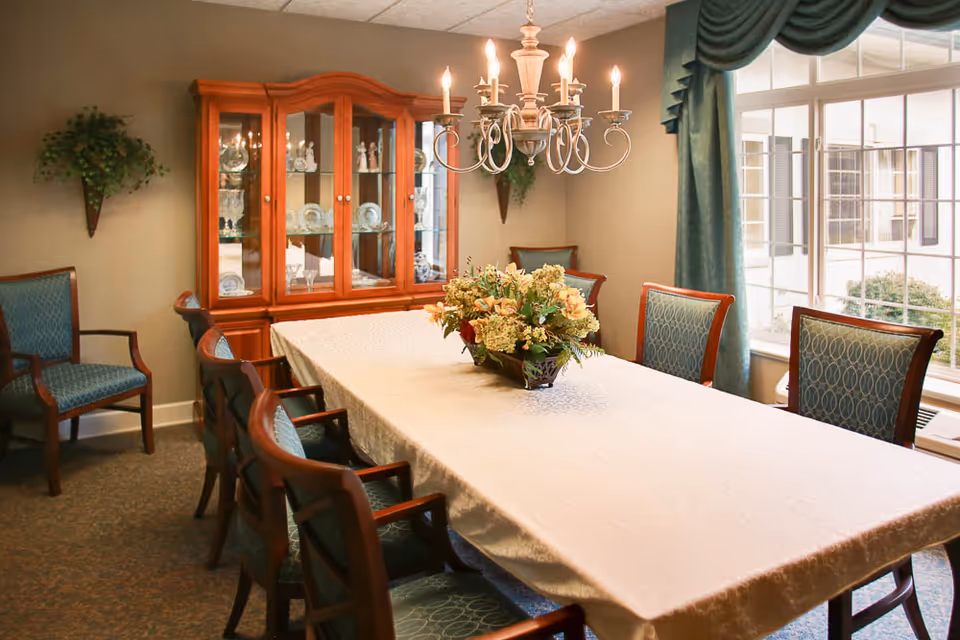 A formal dining room with a long rectangular table covered with a white tablecloth, surrounded by wooden chairs with blue patterned cushions. A floral centerpiece is placed in the middle of the table. A wooden china cabinet with glass doors displaying dishes and glassware is against the wall. A chandelier with lit candles hangs above the table. There is a large window with blue curtains letting in natural light, and a small wall-mounted plant decoration is visible on the wall.