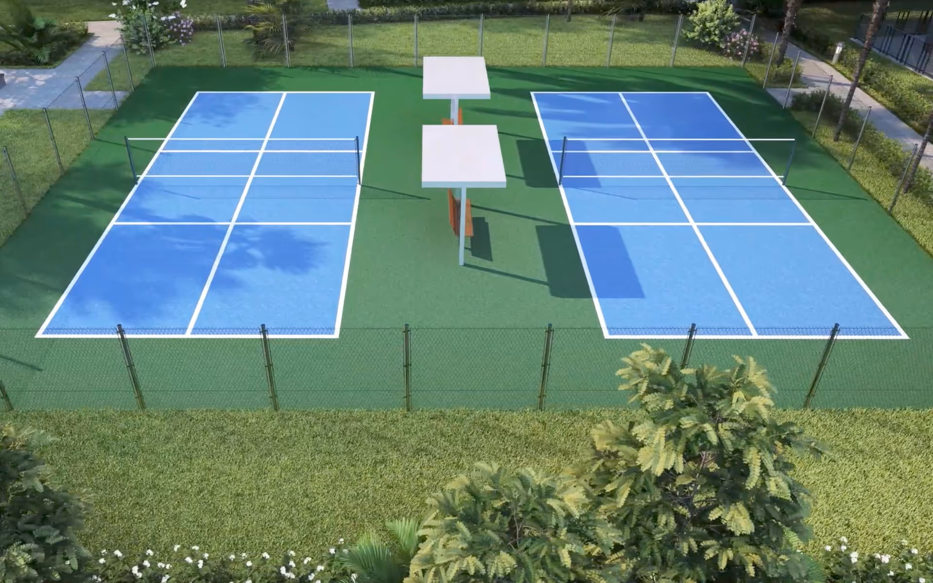 Aerial view of two adjacent blue pickleball courts with white lines, surrounded by green fencing and greenery. There are two shaded benches between the courts, and the area is enclosed by grass and trees.