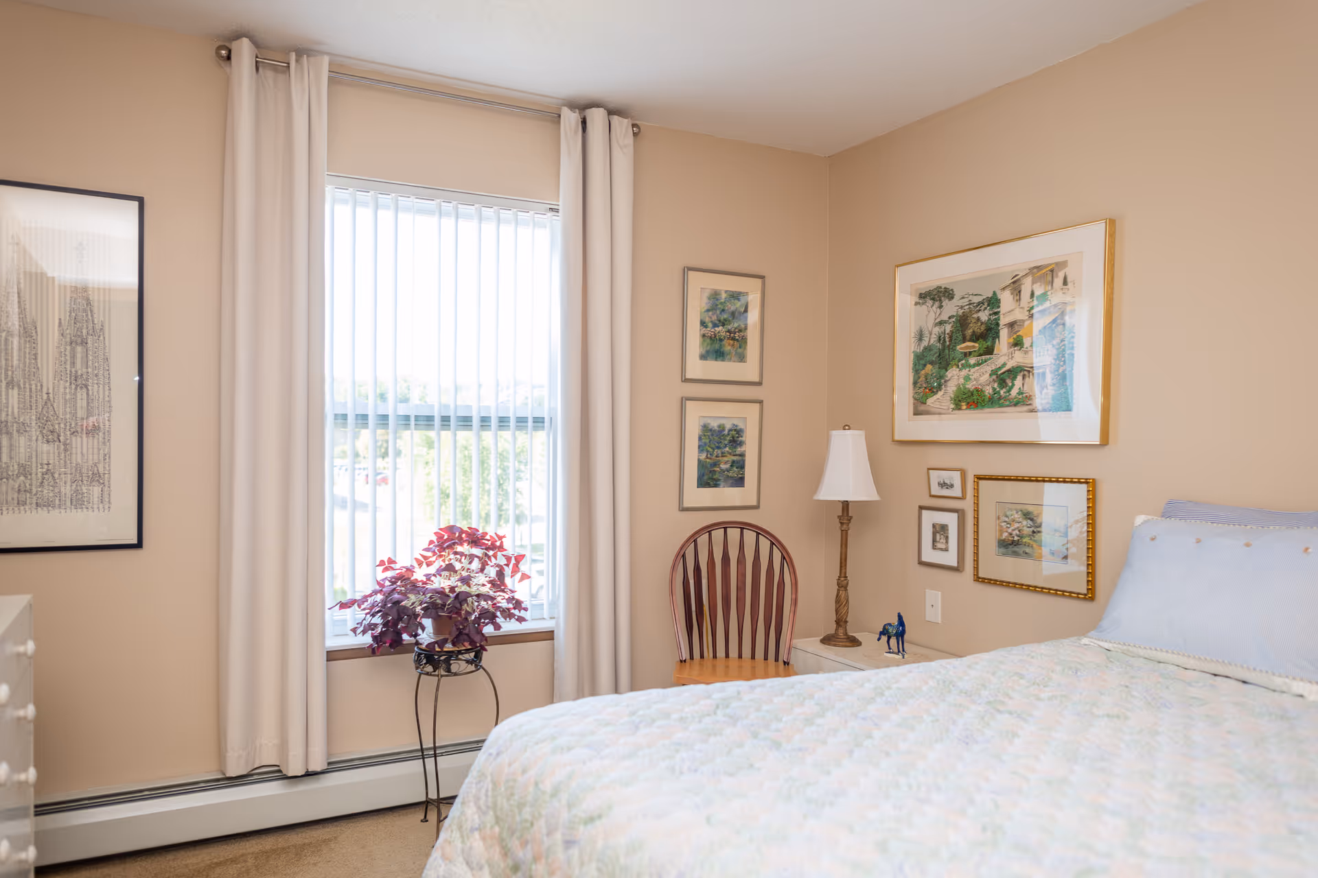 A cozy bedroom with a bed covered in a light-colored quilt, a wooden chair, a small white nightstand with a lamp and decorative items, several framed pictures on beige walls, a window with vertical blinds and white curtains, and a potted plant on a stand near the window.