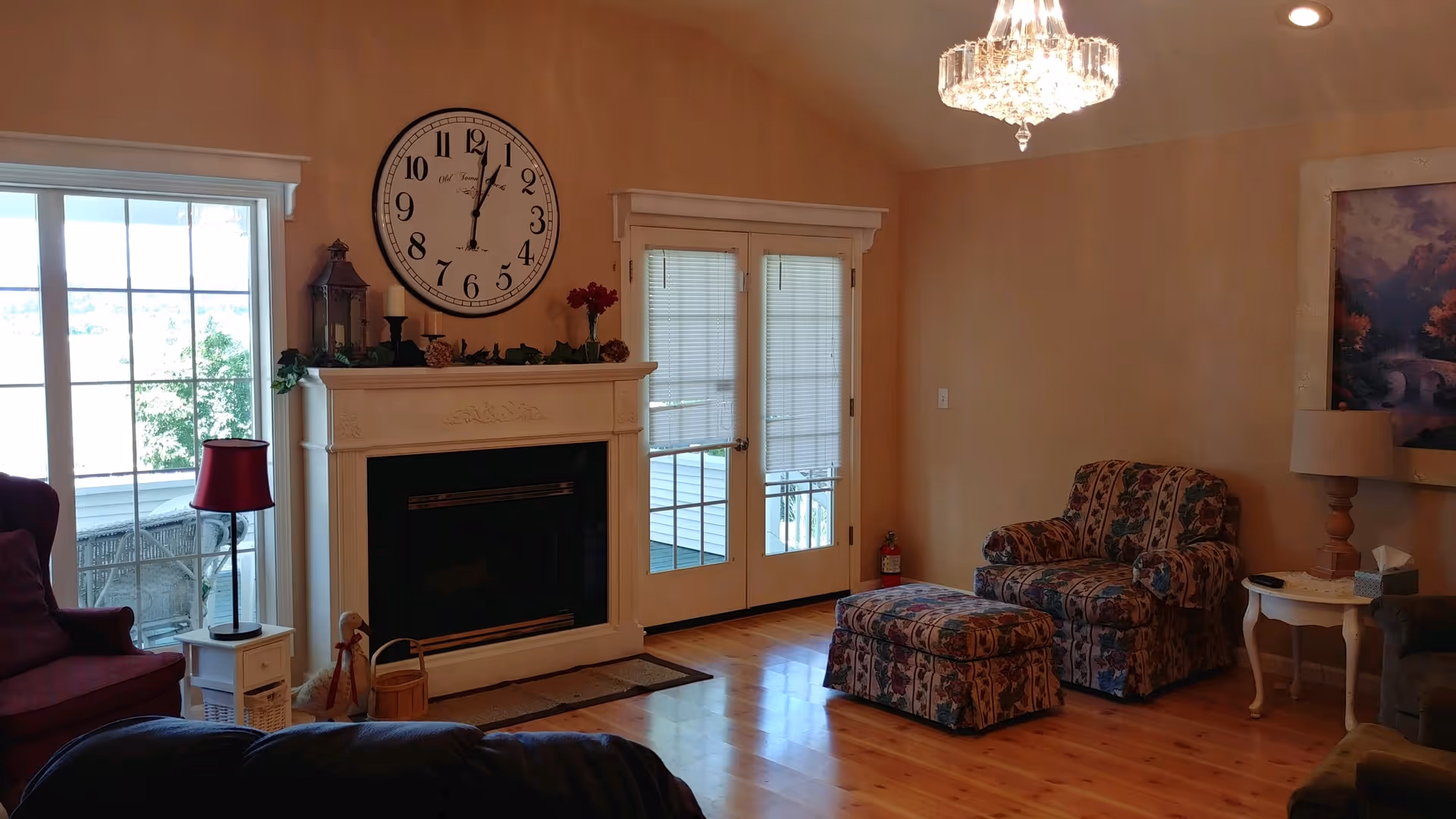 Cozy living room with a fireplace topped by a large wall clock, patterned armchair and ottoman, and French doors to the outside.