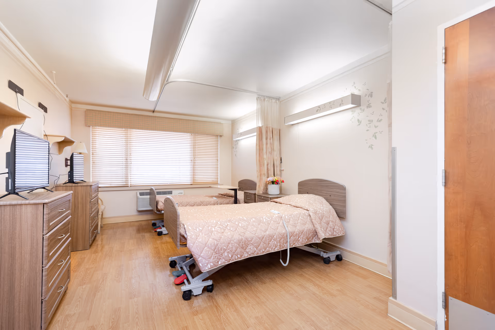 A bright and clean room with two hospital-style beds covered in light pink quilts, a wooden dresser with a TV on top, a window with blinds, and a wooden door. The room has light-colored walls and wood flooring, with a small table and chair near the window.