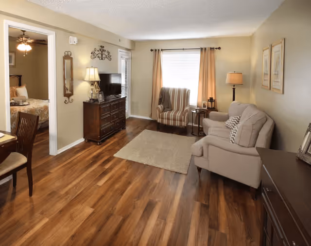 Well-lit living room with a sofa and armchair, TV on a dresser, wood-look floors, and a doorway leading to a bedroom.
