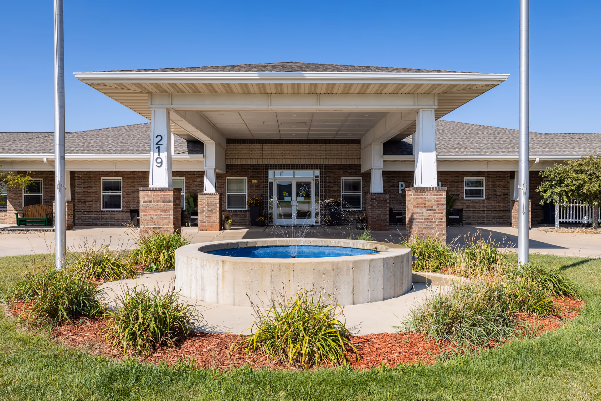 Front entrance of a single-story brick senior living building with a covered drop-off area, flagpoles, and a circular fountain in the foreground.