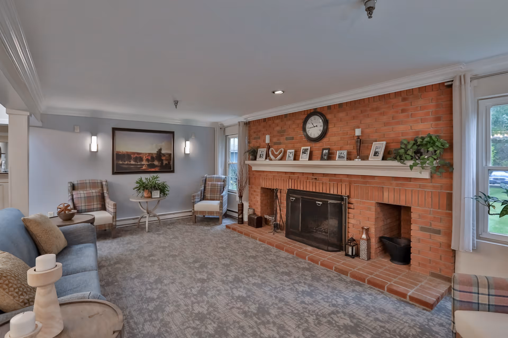 A cozy living room with a large brick fireplace as the focal point. The fireplace mantle is decorated with framed photos, candles, a clock, and a plant. The room has a soft gray carpet, a blue sofa with beige cushions, two plaid armchairs, a small round table with a plant, and wall sconces flanking a landscape painting. Large windows with white curtains allow natural light into the space.