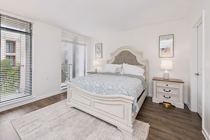A bright bedroom with a large white wooden bed featuring a tufted headboard, covered with a light blue and white patterned comforter and pillows. There are two matching white nightstands on either side of the bed, each with a lamp. The room has wooden flooring and a light-colored area rug. Large windows with white blinds allow natural light to fill the space.