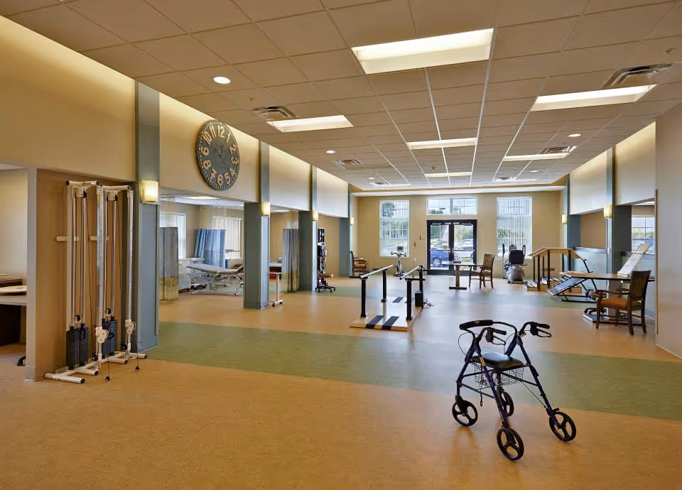 A spacious rehabilitation or physical therapy room with exercise equipment, parallel bars for walking practice, a walker, and several chairs. The room has large windows letting in natural light and a large clock on the wall.