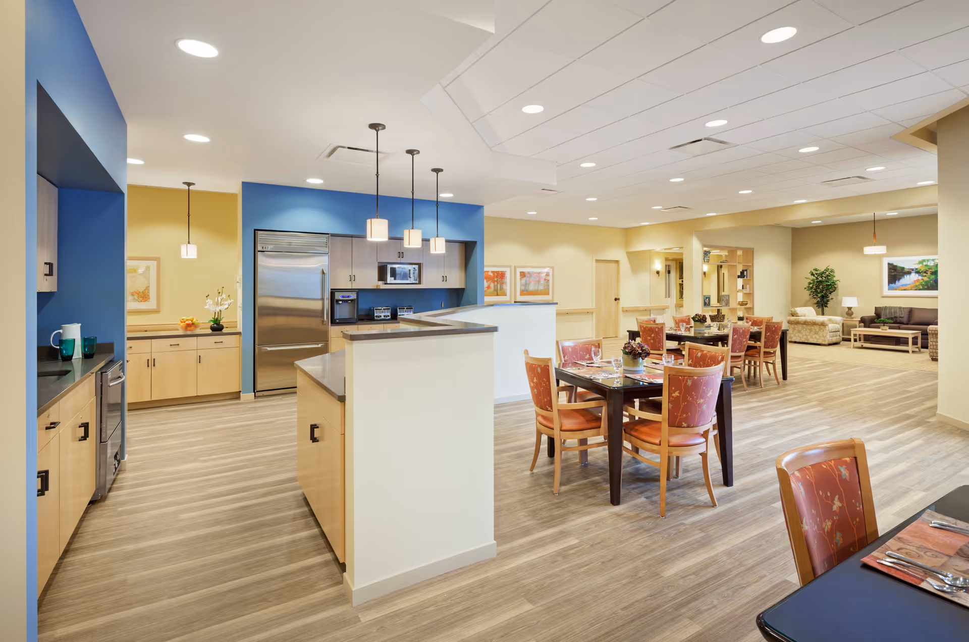 A spacious and well-lit interior area featuring a modern kitchen with stainless steel appliances and light wood cabinetry on the left. Adjacent to the kitchen is a dining area with multiple tables and chairs arranged neatly. The room has light-colored walls with blue accent walls around the kitchen area, wood-look flooring, and ceiling lights. In the background, there is a comfortable seating area with sofas and a coffee table, along with framed artwork and a potted plant.