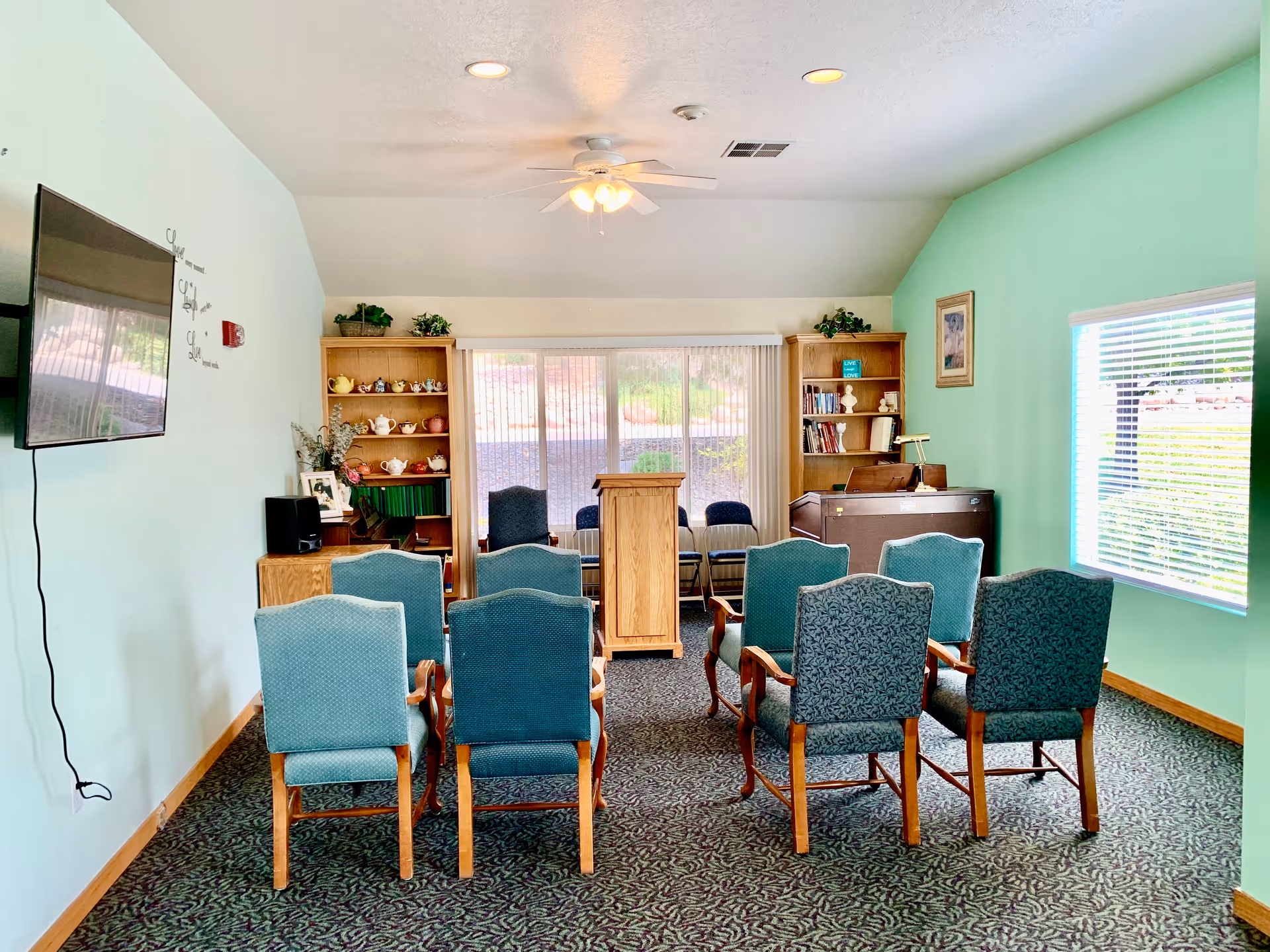 Small community room with rows of upholstered chairs facing a wooden podium, bookshelves, a piano and a wall-mounted TV.