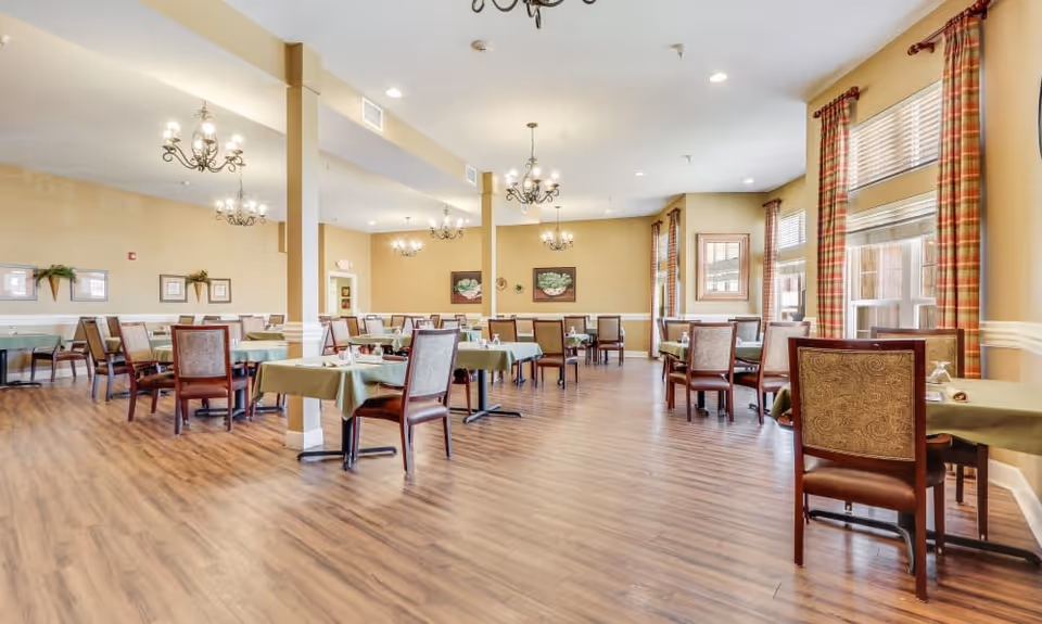A spacious dining room with multiple tables covered in green tablecloths, each surrounded by wooden chairs with cushioned seats and backs. The room features wood flooring, beige walls, several chandeliers hanging from the ceiling, and large windows with plaid curtains allowing natural light to fill the space. Decorative framed artwork and plants adorn the walls.