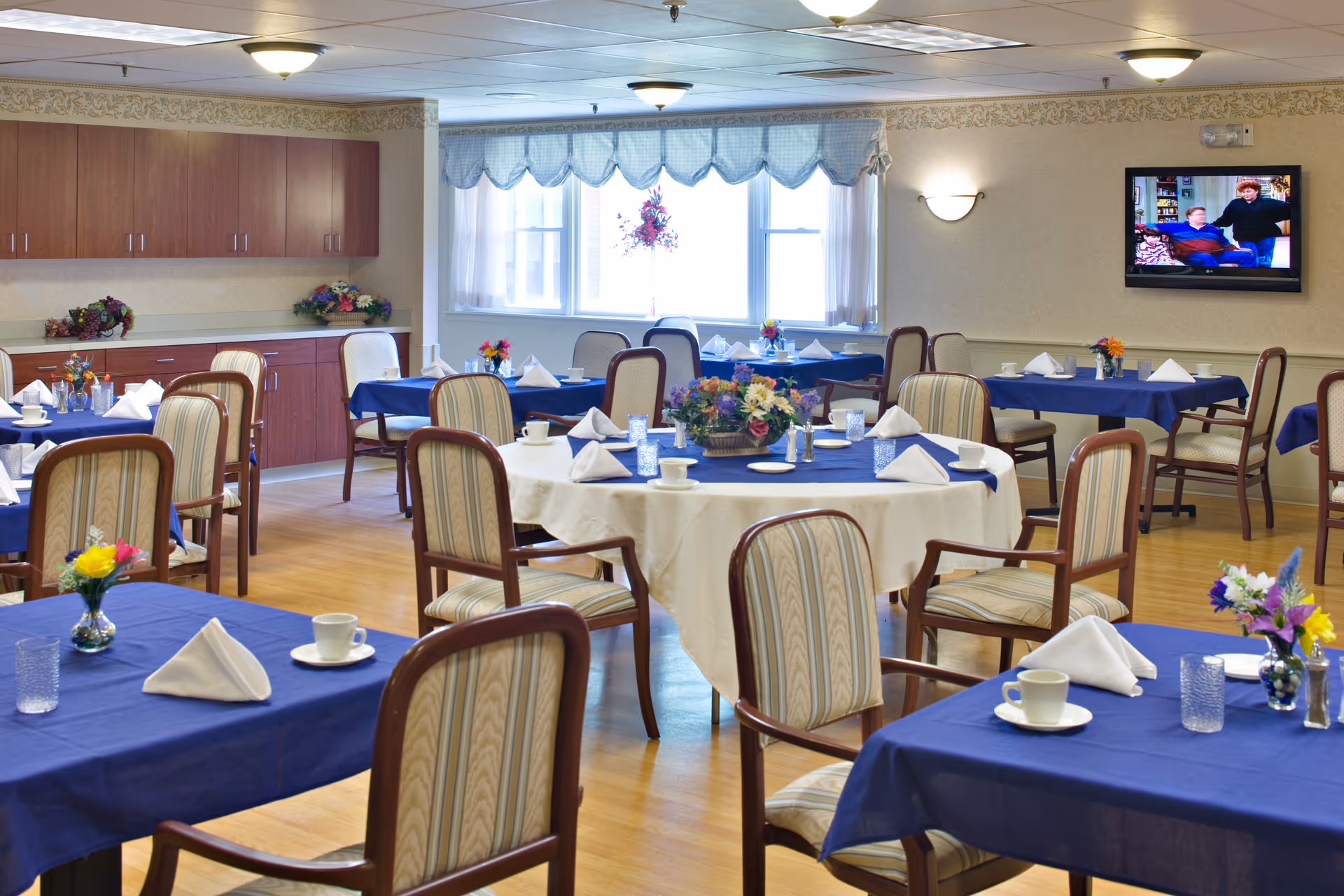 A bright dining room with multiple tables covered in blue and white tablecloths, each set with white napkins, cups, and small flower arrangements. The room has wooden chairs with cushioned seats and backs, a large window with a blue valance, and a wall-mounted TV showing a scene with two people. There are cabinets along one wall with decorative flowers on top.