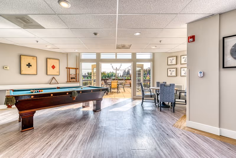 Bright senior living common area with a pool table in the foreground, a table with four chairs to the right, and large glass doors leading to an outdoor patio with additional seating and greenery.