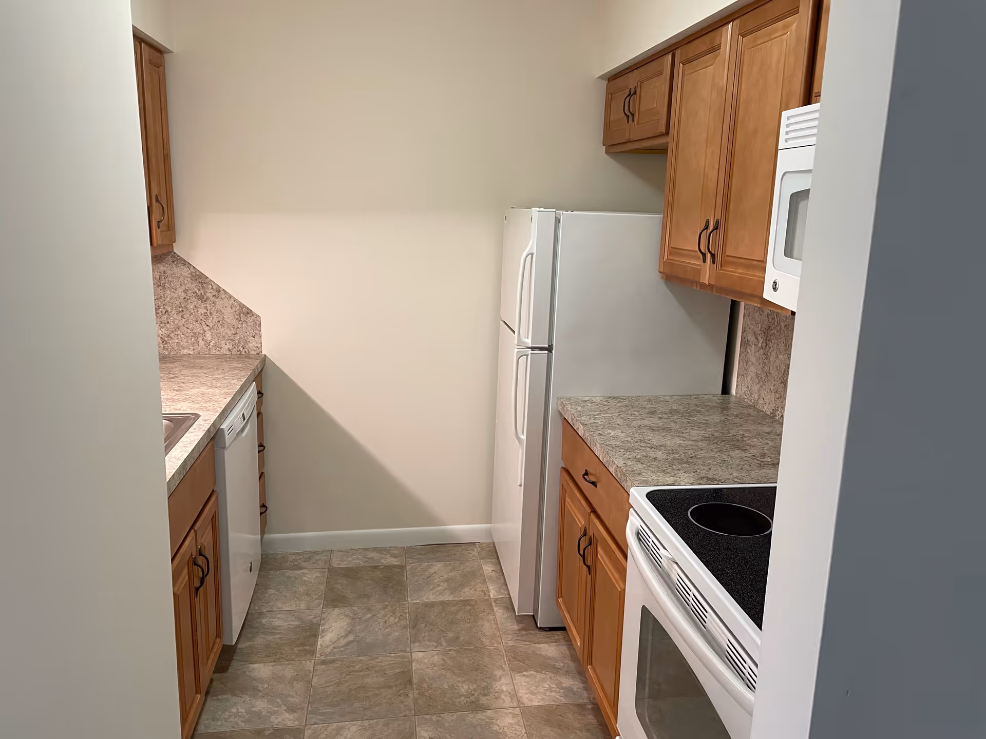 A small kitchen with wooden cabinets, a white refrigerator, a white microwave, a white electric stove, a dishwasher, and a sink. The countertops and backsplash have a stone-like pattern, and the floor is covered with square tiles in neutral tones.