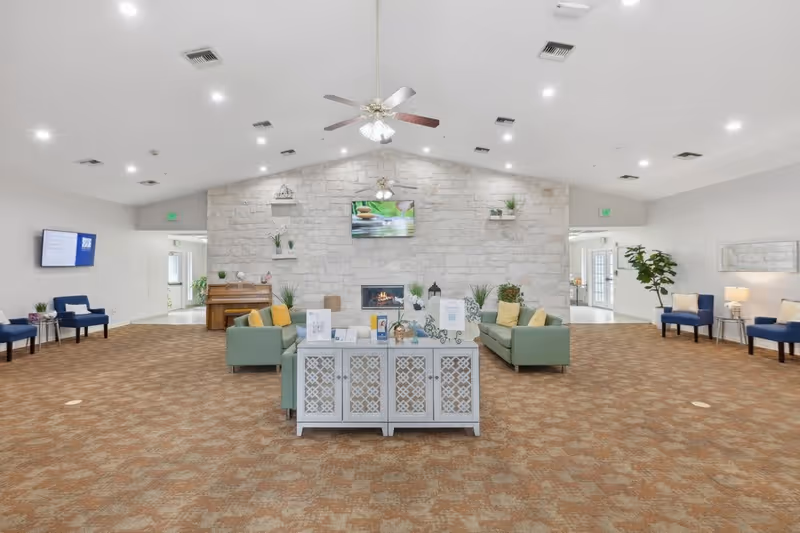 Spacious senior living facility common area with beige patterned carpet, two green sofas with yellow and green pillows facing a stone wall with a fireplace and a mounted TV above it. Ceiling fans with lights hang from the vaulted ceiling. Blue chairs with small side tables and lamps are arranged along the walls. A white decorative cabinet with brochures and decor is placed in the center. Potted plants and a piano are visible in the background.
