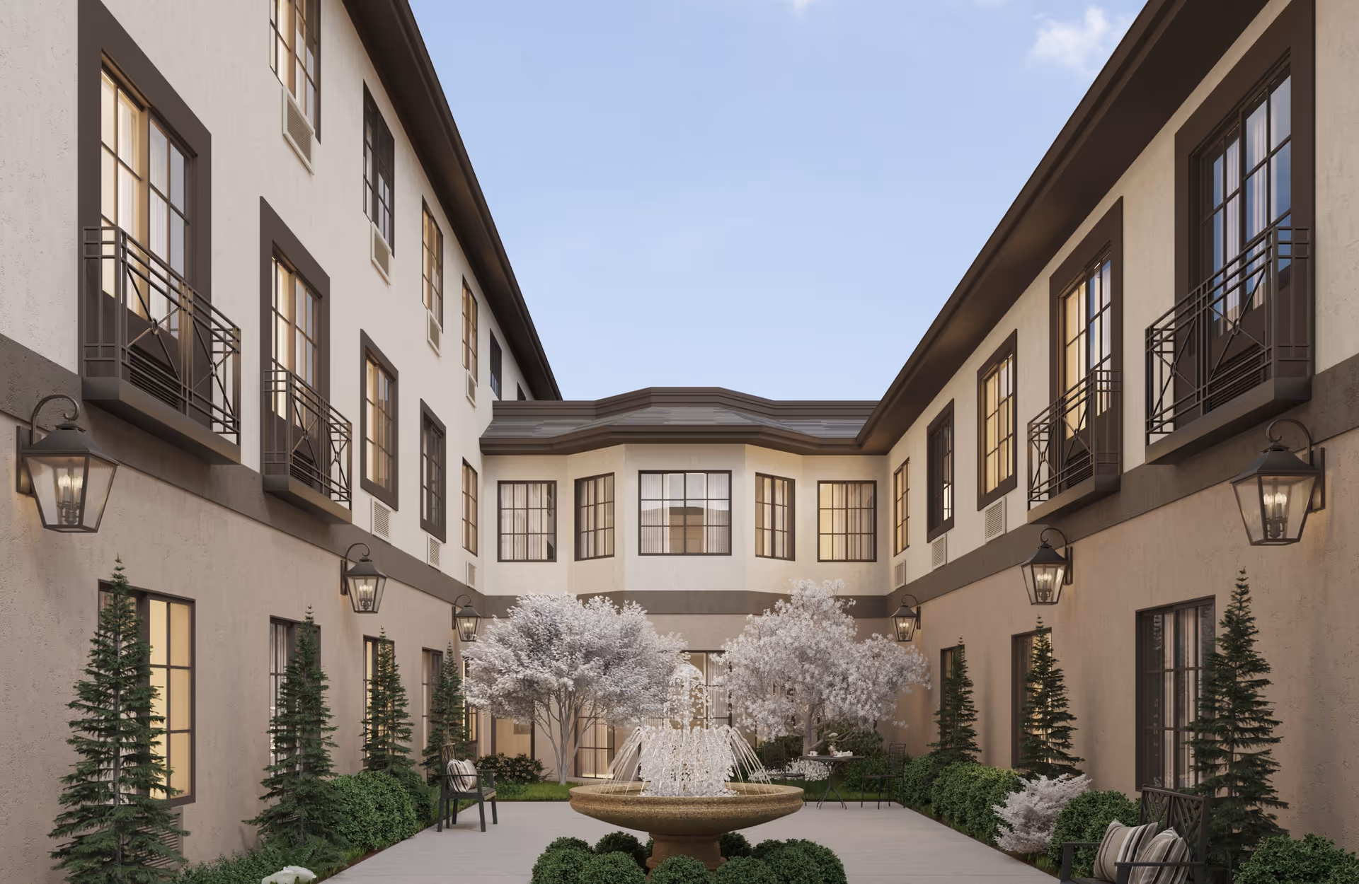 Outdoor courtyard area of a senior living facility with a central water fountain, surrounded by neatly trimmed bushes, small trees with white blossoms, benches, and wall-mounted lanterns on the beige building walls with multiple windows.