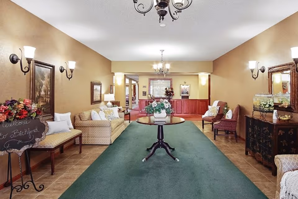 A warmly lit interior common area with beige walls and a green carpet runner. The space features a round wooden table with a floral arrangement in the center, sofas and chairs with cushions along the walls, framed paintings, wall sconces, and chandeliers hanging from the ceiling. There is a decorative blackboard sign with flowers on the left side and a wooden cabinet with a mirror and water dispensers on the right.