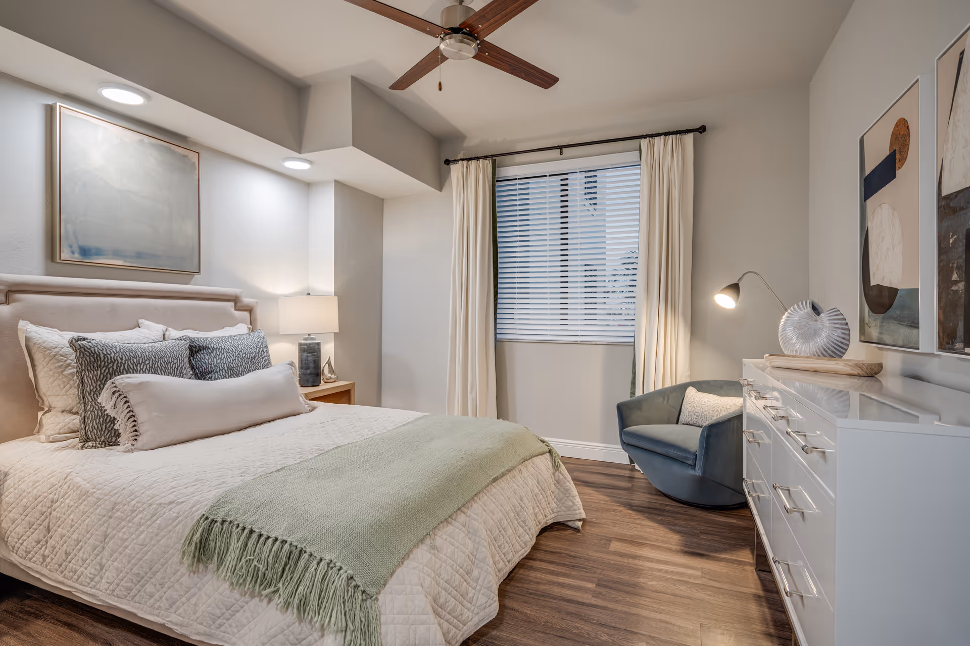 A neatly arranged bedroom with a bed featuring multiple pillows and a light green throw blanket. There is a wooden nightstand with a lamp beside the bed, a ceiling fan above, a window with white blinds and cream curtains, a blue armchair with a pillow, and a white dresser with decorative items and abstract artwork on the wall.