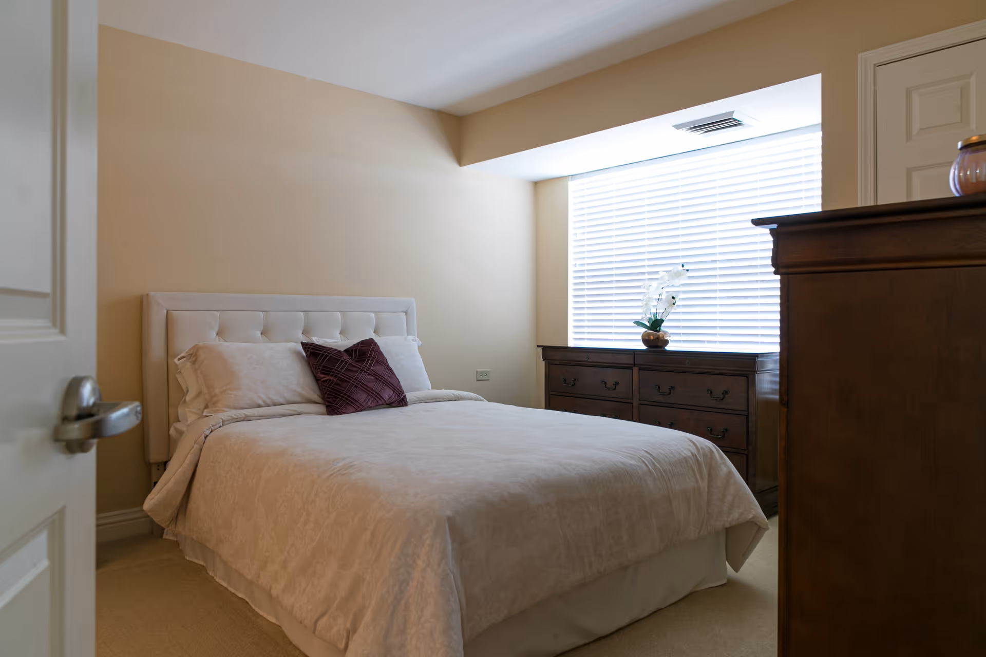 A bedroom with a neatly made bed featuring a white tufted headboard, white bedding, and a dark purple decorative pillow. There is a wooden dresser with drawers beneath a window with white blinds, and another wooden piece of furniture partially visible on the right side.