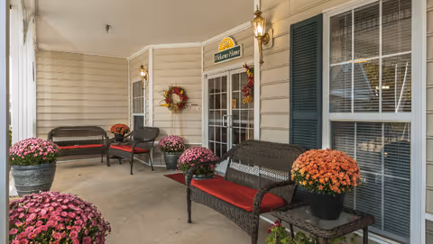 Covered front porch with wicker benches topped with red cushions, potted chrysanthemums, decorative wreaths, and a glass double-door entrance.