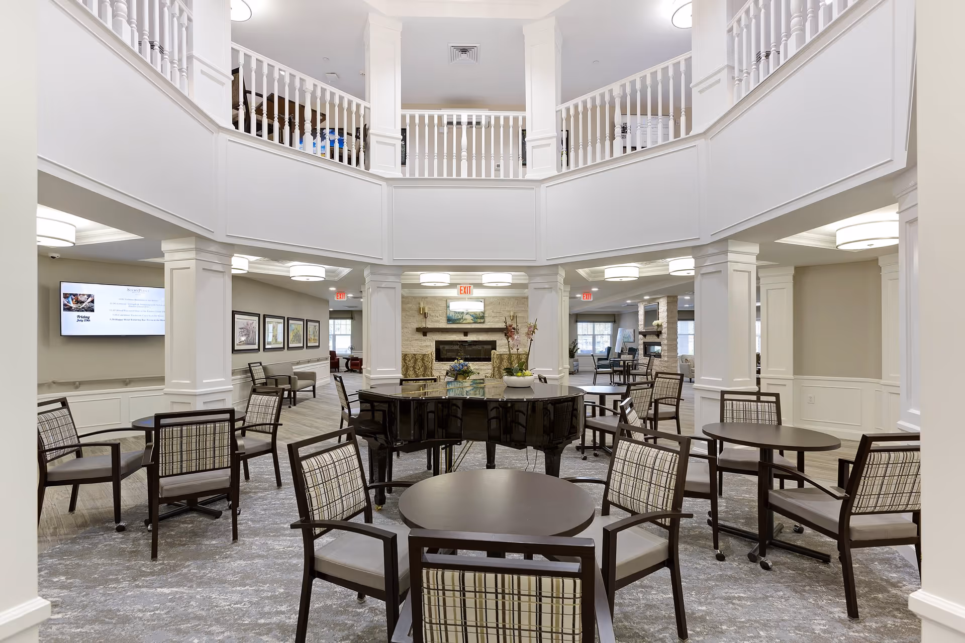 Spacious two-story common lounge with round tables, plaid-upholstered chairs, a grand piano and a fireplace beneath an upper balcony.