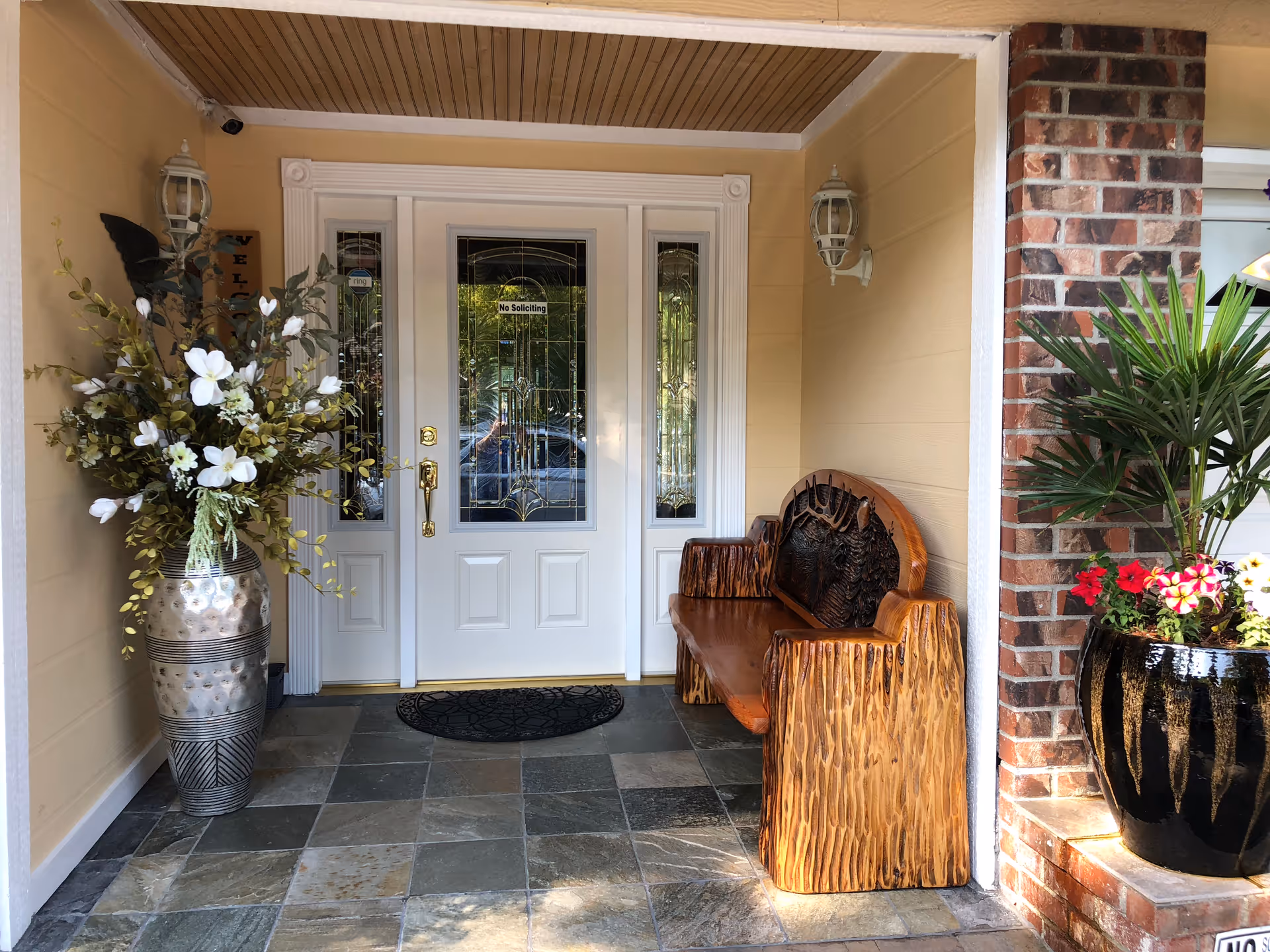 Covered entrance area with a white double door featuring decorative glass panels. To the left, there is a large silver vase with white and green flowers. To the right, a carved wooden bench is placed against the wall. A black doormat is in front of the door, and a black planter with green and red plants sits on a brick ledge to the right.