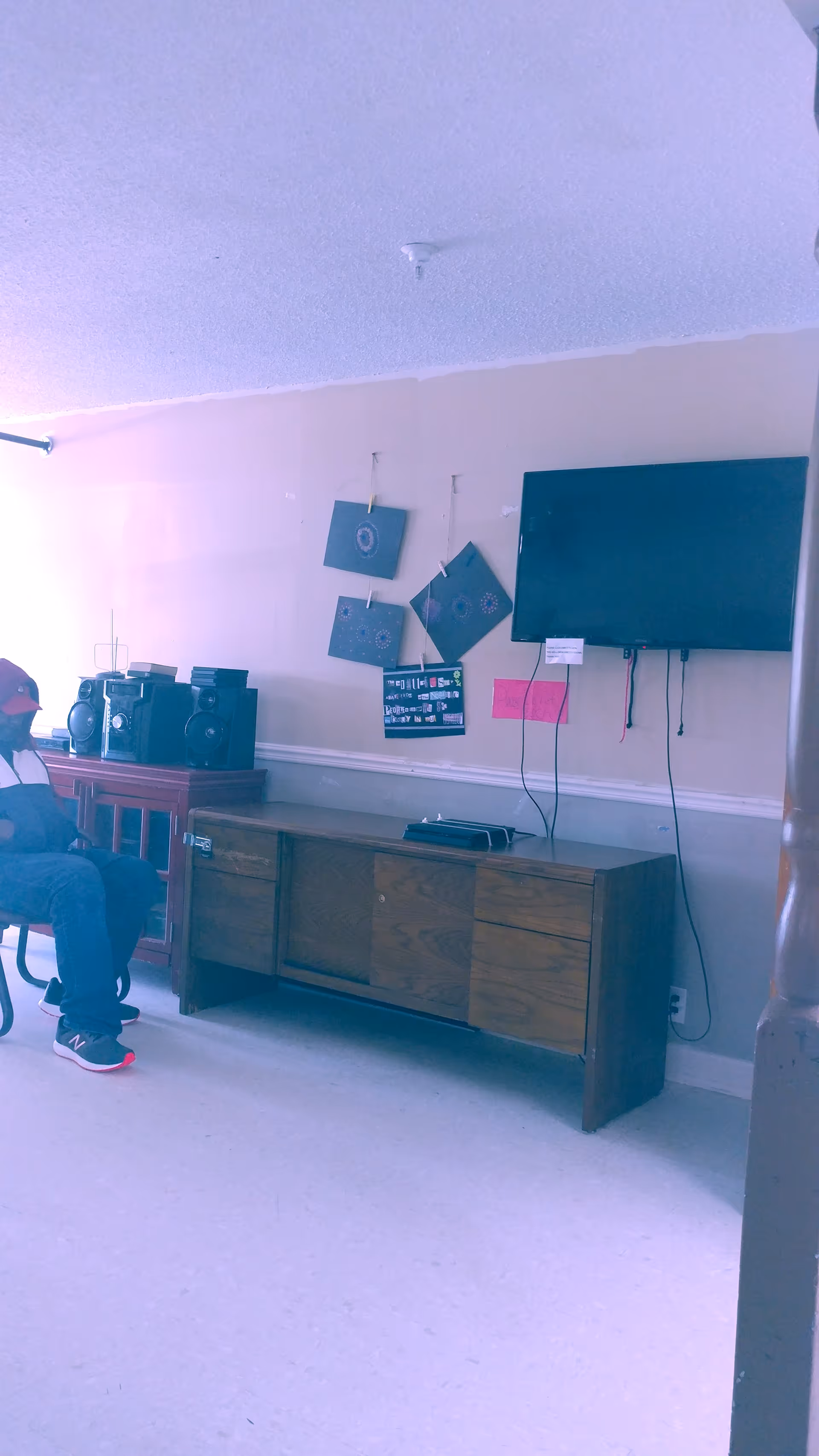 A common room with a wall-mounted TV above a wooden cabinet, a stereo, wall decorations, and a seated person at the left edge.