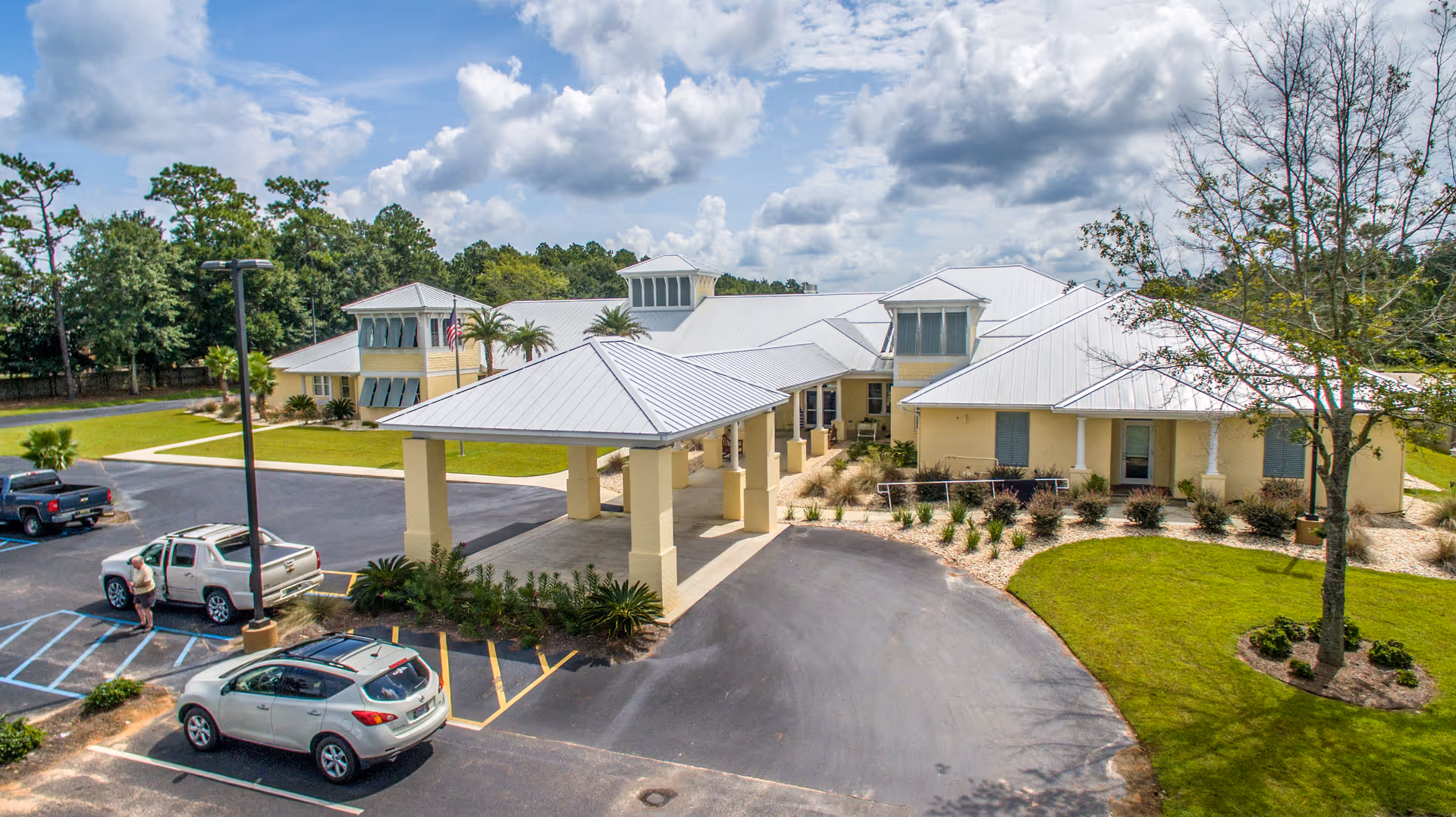 Aerial view of a single-story senior living facility with a covered entrance, surrounded by a parking lot with several cars and landscaped greenery including trees and shrubs under a partly cloudy sky.