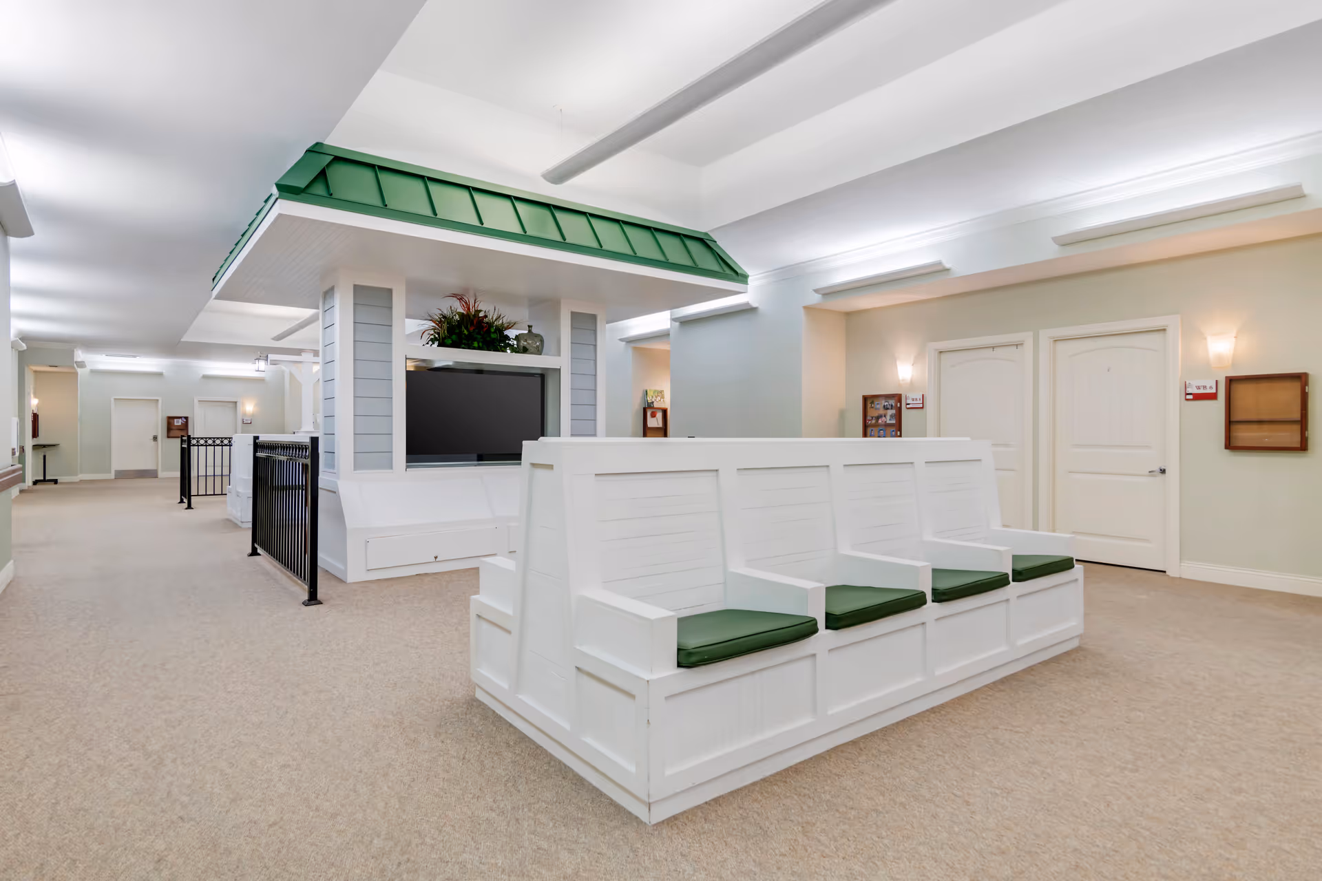 Interior view of a senior living facility hallway with white wooden bench seating featuring green cushions. The area includes a large TV mounted under a green roof-like structure with decorative plants on top. The walls are light-colored with doors and wall-mounted lights, creating a clean and welcoming atmosphere.