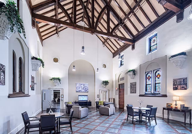 Spacious common area with high vaulted wooden ceiling and exposed beams, featuring several seating arrangements including armchairs and tables with chairs. The room has large stained glass windows, decorative wall sconces with greenery, and framed artwork on white walls. The floor is tiled with a checkered pattern, and pendant lights hang from the ceiling.