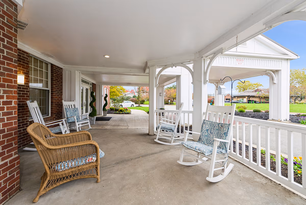 Covered outdoor porch area with white rocking chairs and a wicker chair with cushions, adjacent to a brick building. The porch overlooks a landscaped garden and parking area with trees and a clear sky in the background.