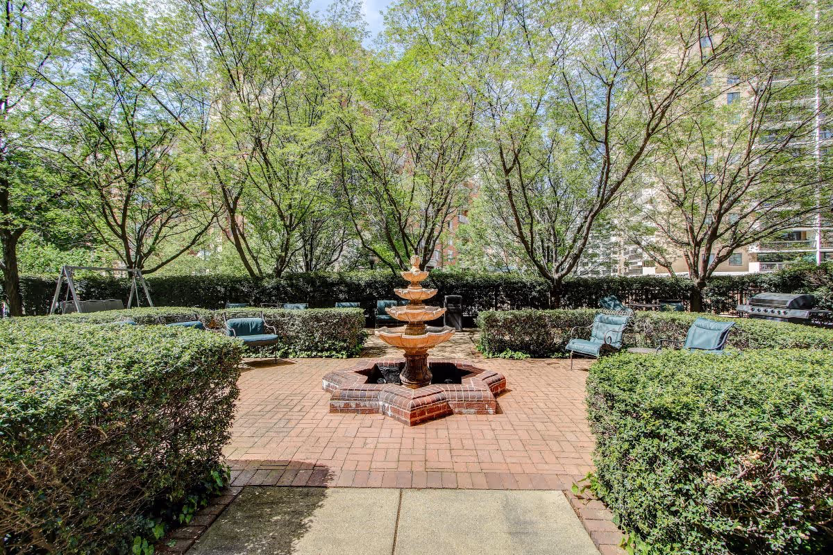 Outdoor patio area with a multi-tiered brick fountain in the center, surrounded by green bushes and trees. Several cushioned chairs are arranged around the fountain, and a barbecue grill is visible in the background.