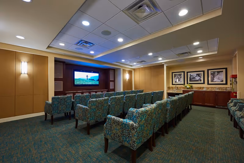 A small theater room with rows of patterned upholstered chairs facing a large flat-screen TV mounted on a wooden panel wall. The room has recessed lighting in the ceiling, beige walls with wall sconces, and framed black-and-white photographs on one side above a wooden cabinet with a granite countertop. A popcorn machine is visible in the corner.