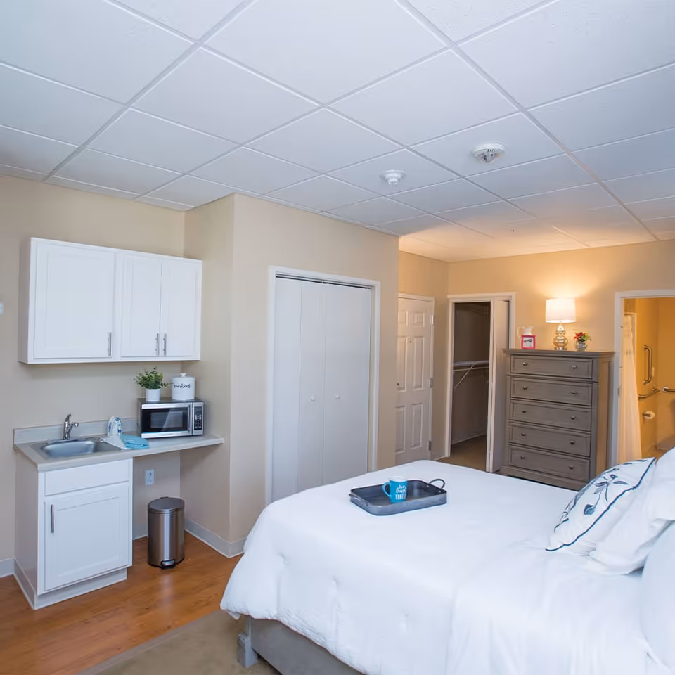A bright and clean assisted living bedroom featuring a large bed with white bedding and decorative pillows. To the left, there is a small kitchenette area with a sink, countertop, microwave, and white cabinets. A trash can is placed underneath the counter. In the background, there is a tall dresser with a lamp and small decorative items on top, an open closet door, and a bathroom entrance with grab bars visible. The room has beige walls and a white tiled ceiling.
