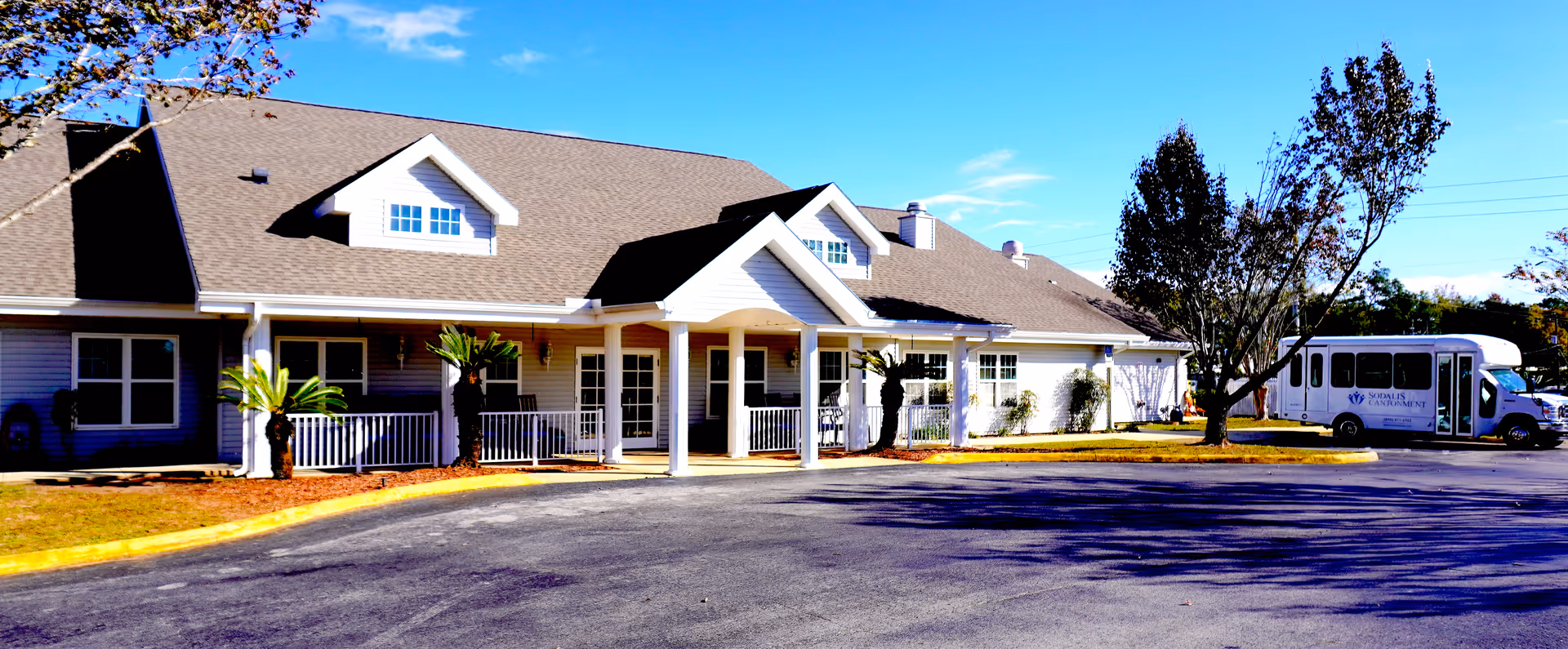 Exterior view of a single-story senior living facility building with a covered entrance, several windows, small palm trees, and a parking area with a shuttle bus parked on the right side under a clear blue sky.